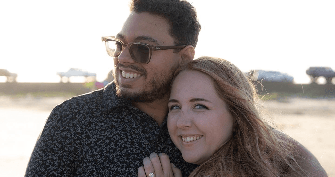 Jess and Nick are standing together at the beach. Jess is looking towards the camera, smiling.